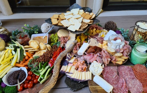 A vibrant charcuterie board with meats, cheeses, crackers, veggies, dips, and decorative greenery on a wooden table.