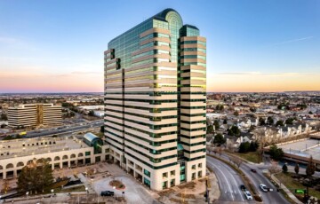 Aerial view of a tall glass and stone office building with a curved top, set against a cityscape at sunset.