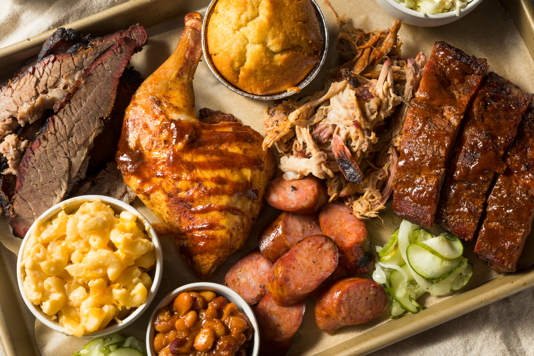 A Denver BBQ catering platter with brisket, chicken, ribs, sausages, corn muffin, mac and cheese, beans, and pickles.