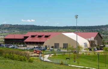 Community center with a red roof, adjacent sports field, and parking lot on a sunny day.