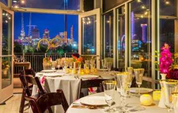 Elegant dining setup with round tables, glassware, and flower arrangements; city skyline and lights visible at night.