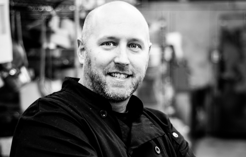 A man in a chef's uniform, smiling, stands in a kitchen. Image is black and white.