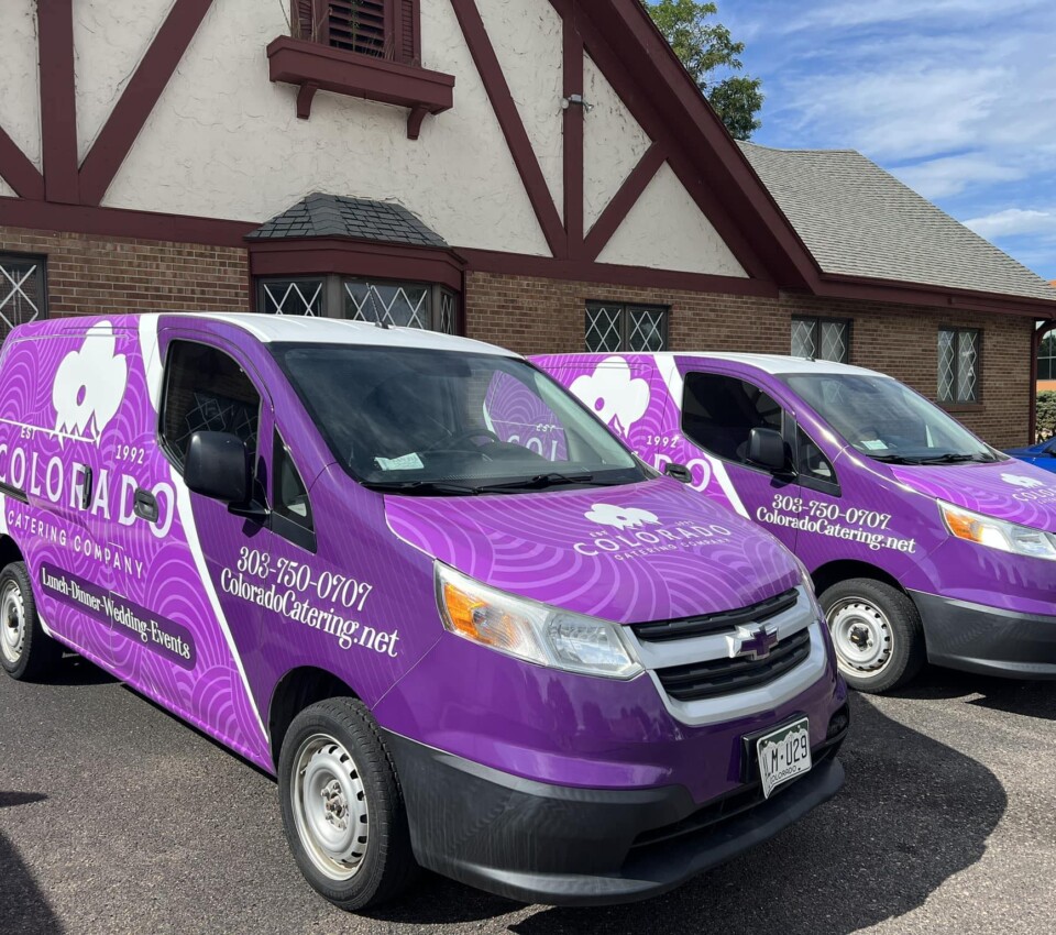 Two purple vans with "Colorado Catering" branding parked in front of a building with a brick exterior.