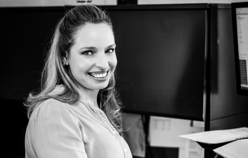 Woman smiling at a desk in an office, with computer monitors in the background, black and white photo.