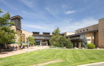 Modern building with glass entrance, surrounded by trees and a grassy lawn under a clear blue sky.