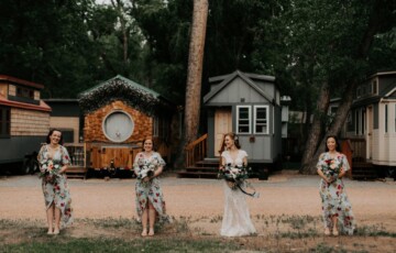 Bride and three bridesmaids holding bouquets, standing in front of tiny houses in a wooded area.