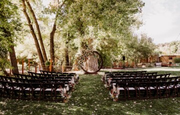 Outdoor wedding ceremony setup with rows of chairs facing a circular floral arch under tall trees.