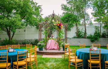 Outdoor event setup with a floral-draped gazebo, tables with blue cloths, and wooden chairs on a lawn.