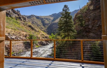 View of a river flowing through a mountain canyon, seen from a bridge with a wooden railing.