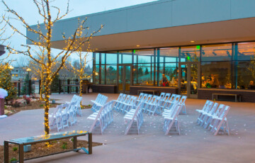 Outdoor wedding venue set up with white chairs, fairy lights, a draped arbor, and glass-walled building in the background.