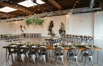 Spacious event hall with wooden tables, chairs, hanging greenery, and decorative plants on white brick walls.