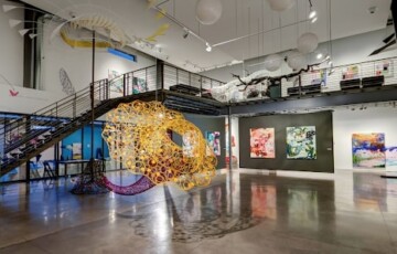 Spacious art gallery with colorful abstract artworks, a metal staircase, and a large circular sculpture centerpiece.