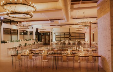 Spacious wedding venue with elegant chandeliers, wooden tables, transparent chairs, and soft candlelight.