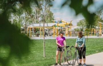 Two people with bicycles stand on a path near a park playground, surrounded by green trees and grass.