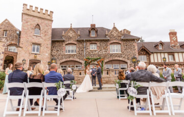 Outdoor wedding ceremony in front of a stone castle-like building, with guests seated on white chairs.