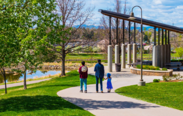 A family walks on a path in a park with a pond, trees, and a pavilion on a sunny day.