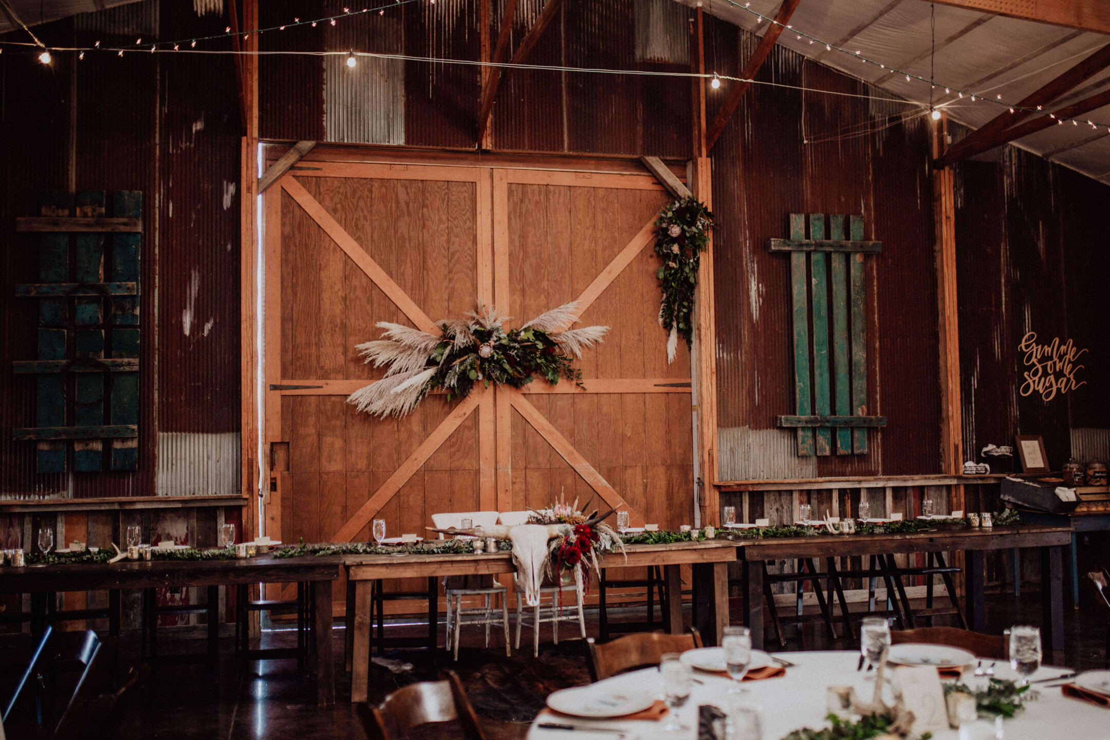 Rustic barn wedding setup with wooden tables, greenery, and floral decor on doors; strings of lights overhead.