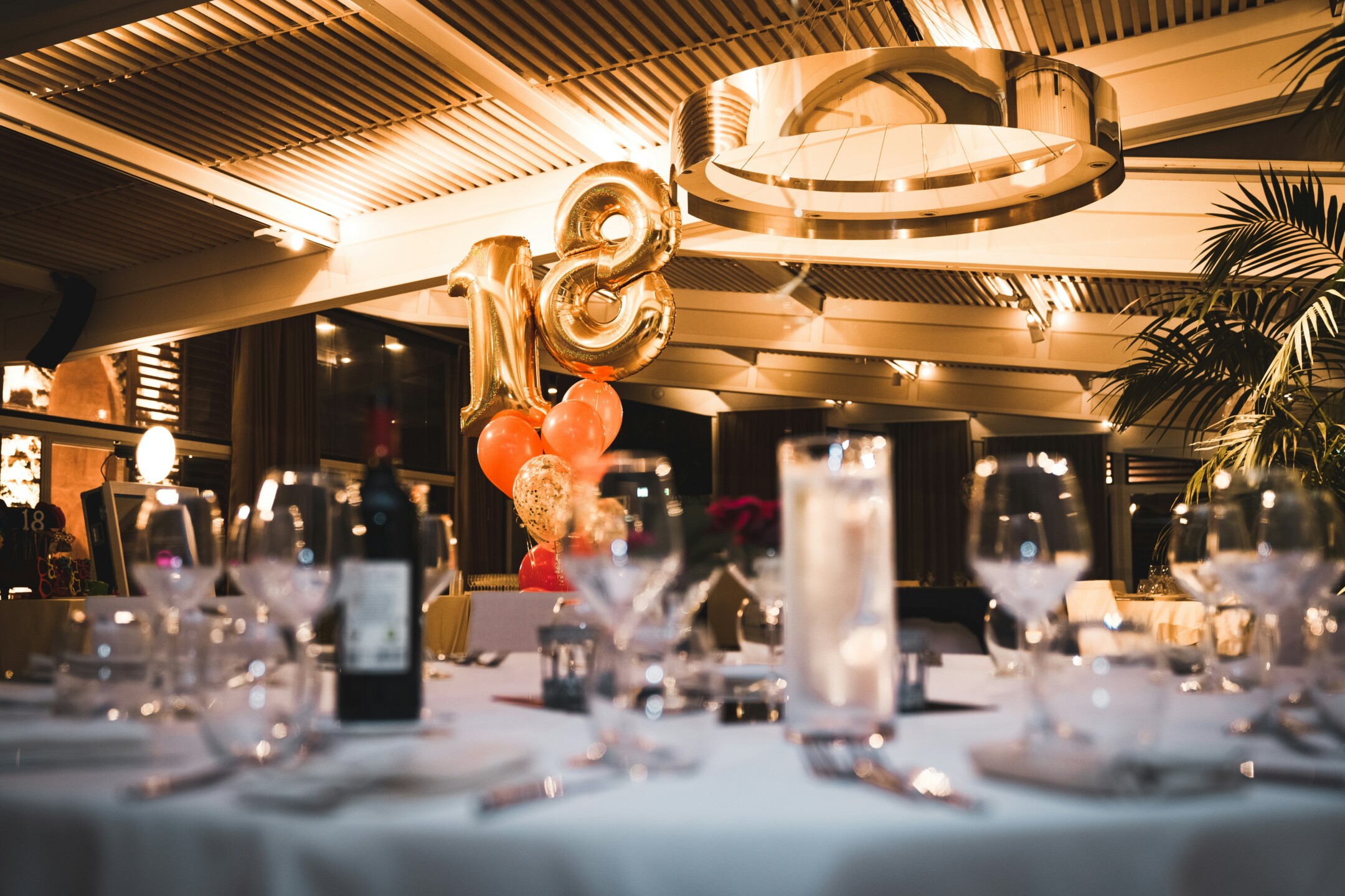 Elegant restaurant with an "18" balloon centerpiece, wine bottle, and glasses on a table, softly lit ambiance.