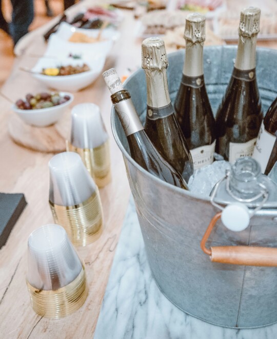 A bucket of champagne bottles on ice, plastic cups, and platters of food on a table.