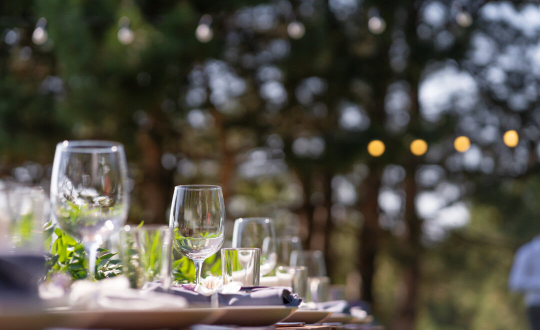 A table set outdoors with empty wine glasses and tableware, surrounded by blurred trees and string lights in the background.