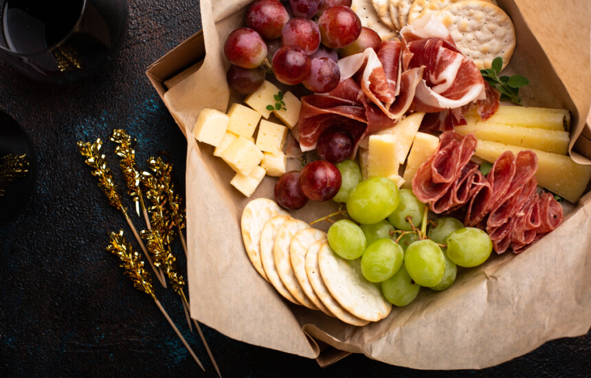 Charcuterie box with grapes, crackers, cheese, and cured meats on dark surface with gold decorative twigs nearby.