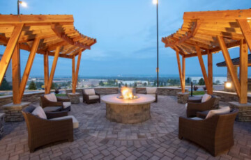 Outdoor seating area with wicker chairs around a lit fire pit, framed by two wooden pergolas against a twilight sky.