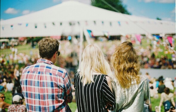 Three people in casual attire stand at an outdoor event, facing a large tent with a crowd and blue sky above.