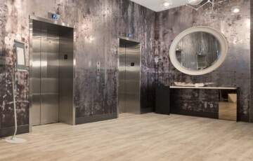 Modern elevator lobby with two elevators, textured walls, large round mirror, and unique ceiling light fixture.