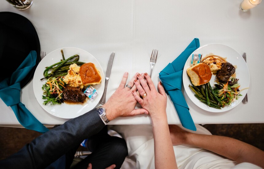 Couple holds hands over a table set with plates of food, including meat, green beans, and rolls, on a white tablecloth.